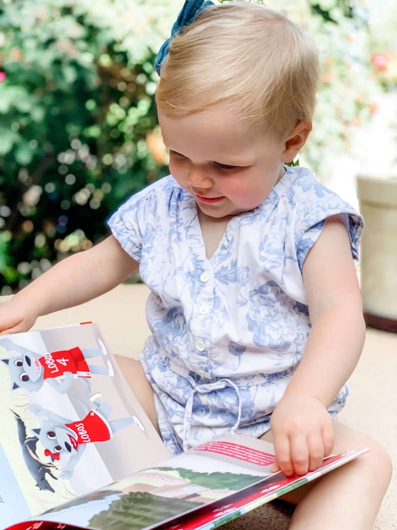 May include: A young child wearing a blue and white floral romper is engrossed in a colorful book. The book features illustrations of cartoon wolves in red jerseys with the word "LOBOS". A blue bow adorns the child's hair.