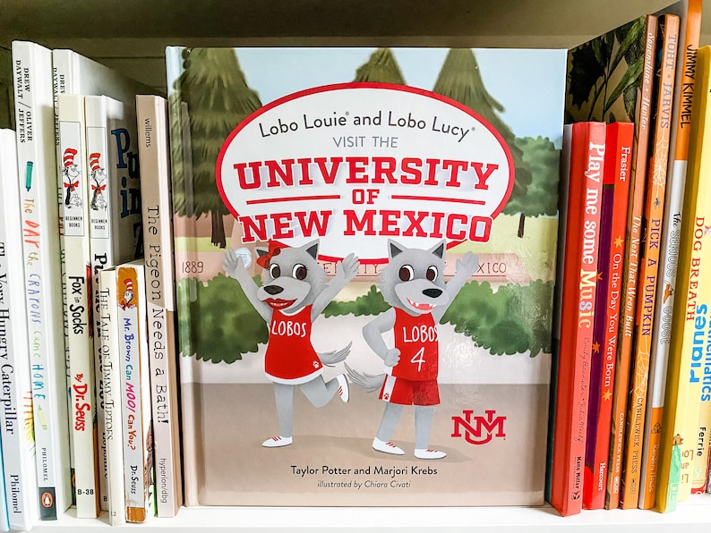 May include: A children's book titled "Lobo Louie and Lobo Lucy Visit the University of New Mexico" is displayed on a shelf with other books. The cover features cartoon wolf characters in red uniforms. The book is surrounded by other colorful books.