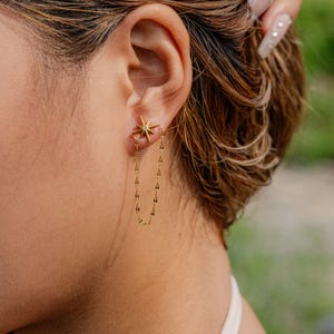 May include: Gold-colored star-shaped earrings with a small gemstone in the center of the star. A delicate chain dangles from the star, creating a stylish and elegant look. The earrings are a beautiful accessory.