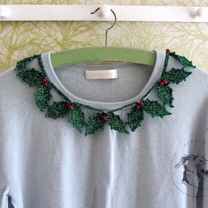 Può includere: Un maglione blu chiaro con un bordo di foglie di agrifoglio all'uncinetto verde e bacche rosse intorno al collo. Il maglione ha un'etichetta che dice "Cachemire".