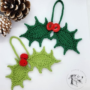 Könnte beinhalten: Drei gehäkelte Stechpalmenblätter-Ornamente mit roten Knopf-Akzenten. Die Ornamente sind grün und rot und perfekt zum Dekorieren eines Weihnachtsbaums.