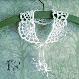 Może przedstawiać: Biały koronkowy kołnierzyk w kształcie pajęczyny z białą, koronkowa pająkiem zwisającym z dołu. Kołnierzyk wisi na zielonym wieszaku na białym tle.