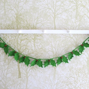 May include: A green crocheted holly garland with red berries hangs on a white wall with a green and white patterned wallpaper.
