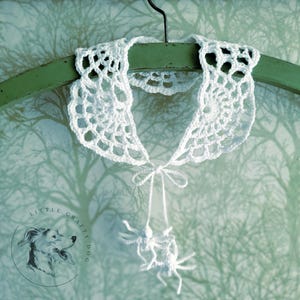 Puede incluir: Collar blanco de ganchillo en forma de telara&ntilde;a con una ara&ntilde;a de ganchillo blanca colgando de la parte inferior. El collar est&aacute; colgado de una percha verde sobre un fondo blanco.