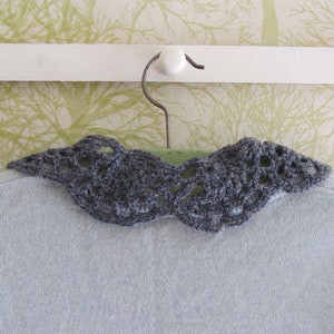 Puede incluir: Un cuello de ganchillo gris con un delicado patr&oacute;n de encaje, colgado de una percha blanca sobre un fondo estampado verde y blanco.