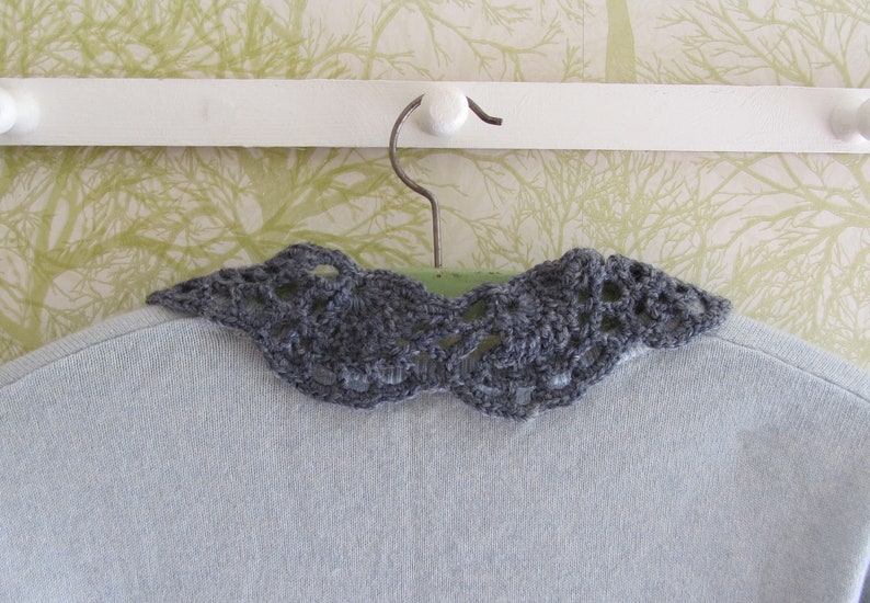 Puede incluir: Un cuello de ganchillo gris con un delicado patr&oacute;n de encaje, colgado de una percha blanca sobre un fondo estampado verde y blanco.