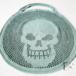 May include: A round, seafoam green crocheted bag with a skull design. The skull is centered on the bag and features openwork eyes and a toothy grin. The bag has a handle for carrying.