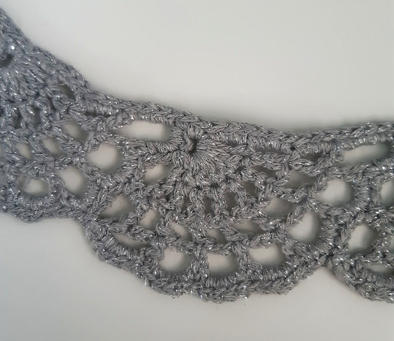 Puede incluir: Un collar de crochet plateado con un dise&ntilde;o delicado y de encaje. El collar est&aacute; hecho de un hilo brillante y tiene un borde festoneado.