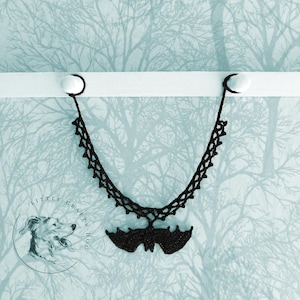 May include: Black crocheted bat necklace hanging on a white rod against a blue and white patterned background.