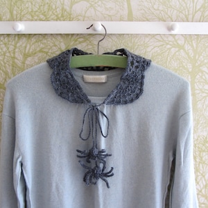 Puede incluir: Un su&eacute;ter azul claro con un cuello de ganchillo gris y una corbata de lazo de ganchillo gris. El cuello tiene un delicado patr&oacute;n de encaje y la corbata de lazo est&aacute; anudada en la parte delantera.