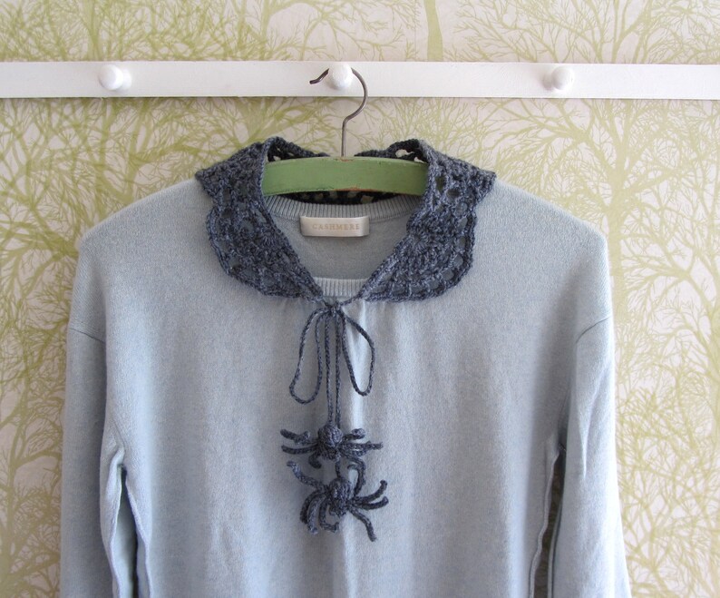 Puede incluir: Un su&eacute;ter azul claro con un cuello de ganchillo gris y una corbata de lazo de ganchillo gris. El cuello tiene un delicado patr&oacute;n de encaje y la corbata de lazo est&aacute; anudada en la parte delantera.