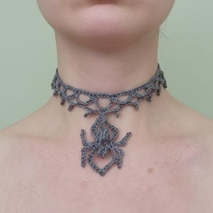 Może przedstawiać: Szary szydełkowy naszyjnik typu choker z wisiorkiem w kształcie pająka. Naszyjnik ma ażurowy wzór wokół szyi i szczegółowy urok pająka wiszący na środku. Pająk ma osiem nóg i teksturowany korpus.