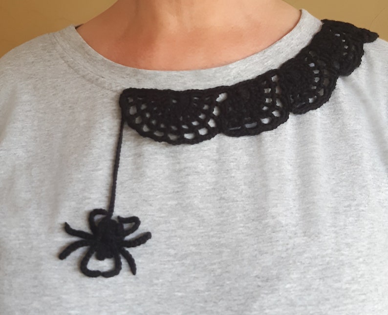 Puede incluir: Una camiseta gris con un dise&ntilde;o de telara&ntilde;a de ganchillo negro alrededor del escote. Una ara&ntilde;a de ganchillo negro cuelga de la telara&ntilde;a.
