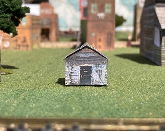 Puede incluir: Una maqueta en miniatura de un cobertizo de madera desgastada con una puerta oscura y un tejado gris, sobre una superficie verde. Otros edificios en miniatura en el fondo sugieren una pequeña ciudad o un diorama.