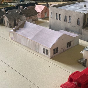 May include: A model train set with a variety of buildings, including a white building with a red roof, a gray building with a flat roof, and a brown building with a pitched roof. There is also a red toy truck in the foreground.