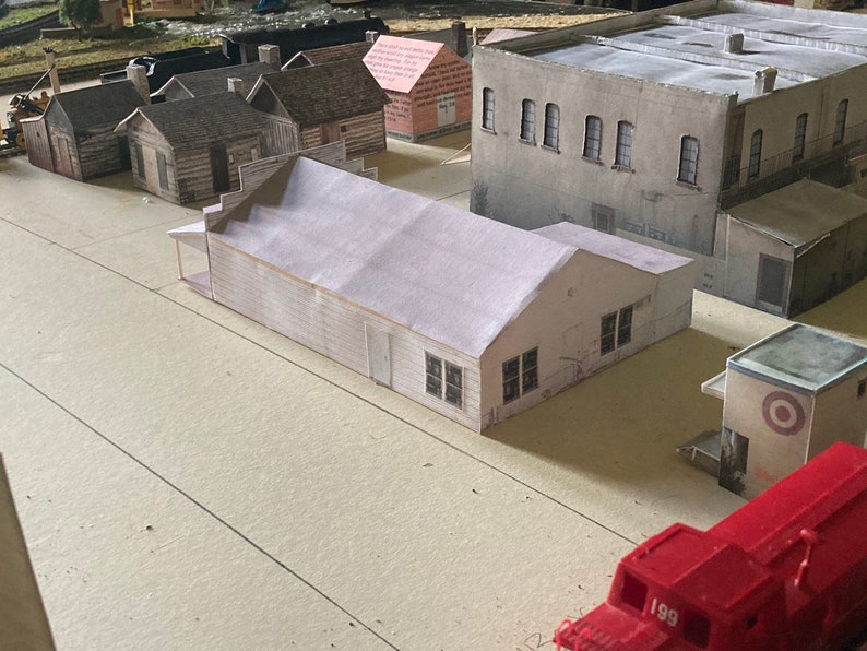 May include: A model train set with a variety of buildings, including a white building with a red roof, a gray building with a flat roof, and a brown building with a pitched roof. There is also a red toy truck in the foreground.