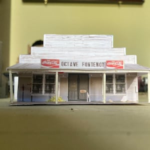 May include: A white paper model of a small store with a red Coca-Cola sign and the text "Octave Fontenot" above the entrance.