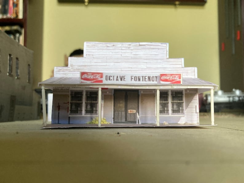 May include: A white paper model of a small store with a red Coca-Cola sign and the text "Octave Fontenot" above the entrance.