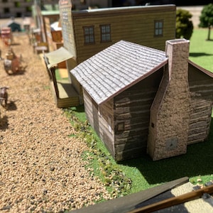 Puede incluir: Una cabaña de troncos en miniatura con un techo de tejas grises y una chimenea de piedra. La cabaña está hecha de madera y tiene un aspecto rústico. La cabaña está situada en una superficie verde cubierta de hierba con un camino de grava en primer plano.