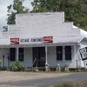 May include: A white building with a sign that reads "Octave Fontenot" and two Coca-Cola signs. There is a sign for LA 103 on the right side of the image.
