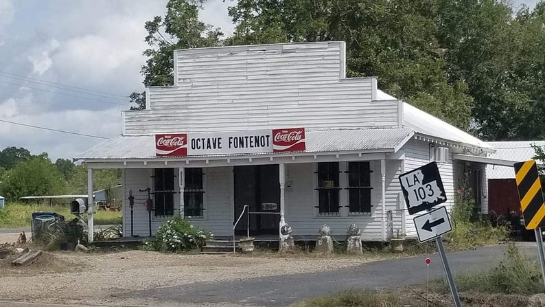 May include: A white building with a sign that reads "Octave Fontenot" and two Coca-Cola signs. There is a sign for LA 103 on the right side of the image.