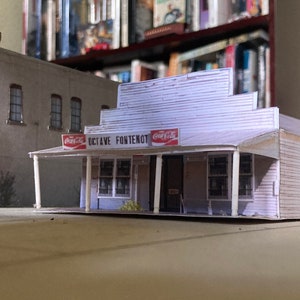 May include: A white paper model of a building with a sign that reads "Octave Fontenot" and two Coca-Cola signs. The building has a porch with white columns and a brown awning.