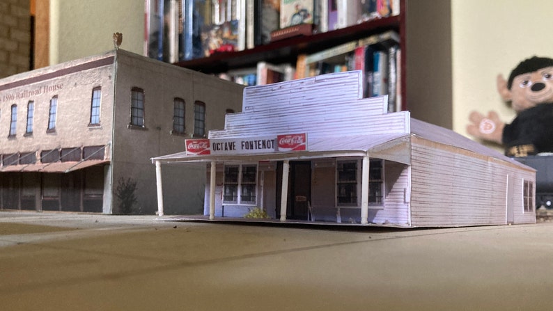 May include: A white paper model of a building with a sign that reads "Octave Fontenot" and two Coca-Cola signs. The building has a porch with white columns and a brown awning.