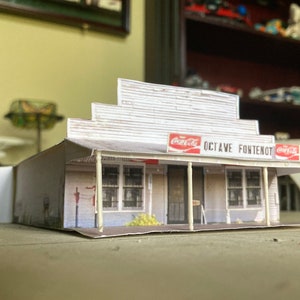 May include: A white paper model of a small building with a Coca-Cola sign and the text "OCTAVE FONTENOT". The building has a porch with white columns and a red roof.