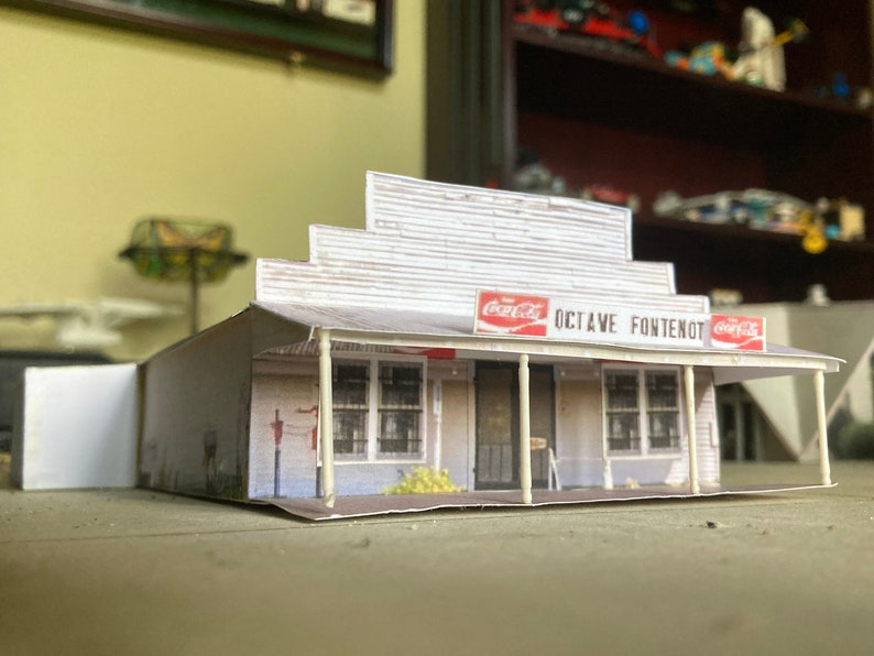 May include: A white paper model of a small building with a Coca-Cola sign and the text "OCTAVE FONTENOT". The building has a porch with white columns and a red roof.