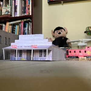 May include: A model of a white building with a Coca-Cola sign and the text "Dixie Ave. Fontenot" on the front. The building is in front of a model train track with a red caboose.