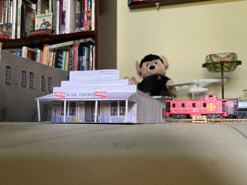 May include: A model of a white building with a Coca-Cola sign and the text "Dixie Ave. Fontenot" on the front. The building is in front of a model train track with a red caboose.
