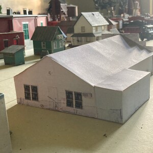 May include: A white model building with a gabled roof and two windows on each side. The building is made of paper and is part of a model train set.