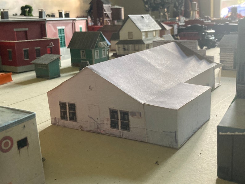 May include: A white model building with a gabled roof and two windows on each side. The building is made of paper and is part of a model train set.