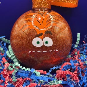 May include: A round, glittery orange Christmas ornament with a cartoon face design. The face has green eyes, a white mouth, and an orange hair design. The ornament has an orange glittery loop for hanging. The ornament is sitting on a bed of red, blue, and green paper shreds.