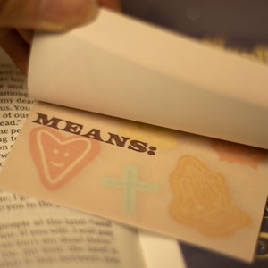 May include: A close-up of a small, brown card with the word "MEANS:" printed in brown letters. The card has a heart with a smiley face drawn on it, a cross, and a hand in a praying position.