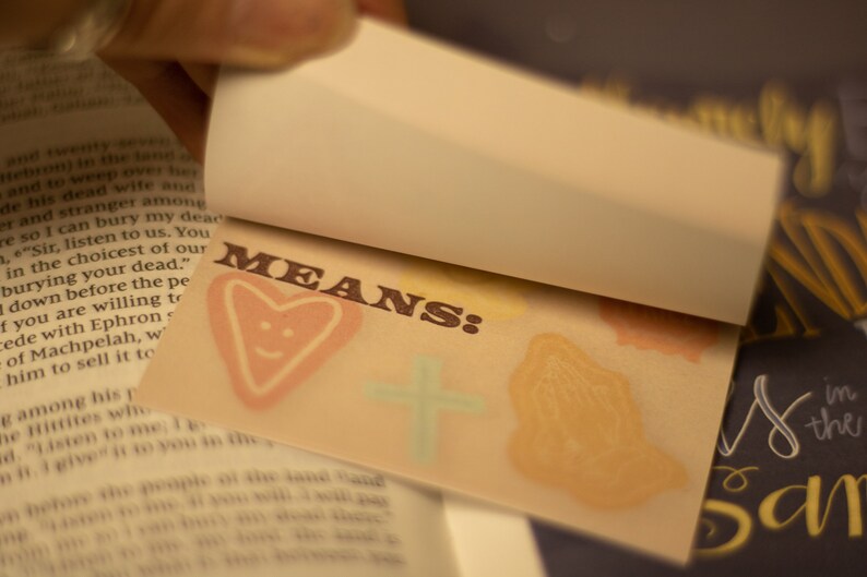May include: A close-up of a small, brown card with the word "MEANS:" printed in brown letters. The card has a heart with a smiley face drawn on it, a cross, and a hand in a praying position.