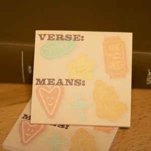 May include: Two beige sticky notes with the words "VERSE: MEANS:" printed on them. The notes are decorated with various hand-drawn images, including hearts, stars, and a ticket with the words "The Price" on it.