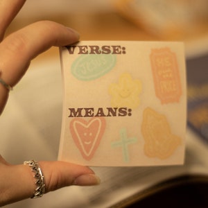 May include: A white card with the words "VERSE:" and "MEANS:" printed in brown. The card has colorful stickers on it, including a heart, a cross, a flower, and a ticket.