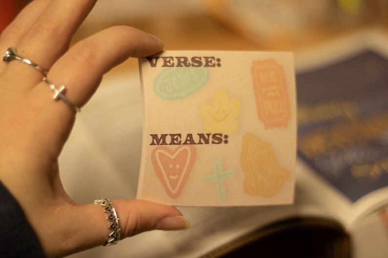 May include: A white card with the words "VERSE:" and "MEANS:" printed in brown. The card has colorful stickers on it, including a heart, a cross, a flower, and a ticket.