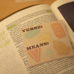 Puede incluir: Primer plano de una página de la Biblia con una nota escrita a mano que dice "The Lord Will Provide". Una nota adhesiva con las palabras "VERSE" y "MEANS" está pegada a la página. La nota adhesiva tiene un corazón, una cruz y otros símbolos.