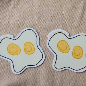 Könnte beinhalten: Zwei weiße Aufkleber in Form von Spiegeleiern mit gelben Smiley-Gesichtern darin. Der Text "Look on the sunny side!" steht auf jedem Aufkleber.