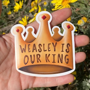 Pode incluir: Um adesivo de coroa dourada com o texto "WEASLEY IS OUR KING" em letras pretas. O adesivo é segurado em uma mão contra um fundo de flores amarelas.
