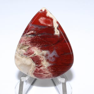 May include: A teardrop-shaped polished gemstone with a striking pattern of deep red, cream, and white hues. The stone's surface displays a mix of textures and colors, creating a unique and natural aesthetic. The gemstone is displayed on a clear stand.