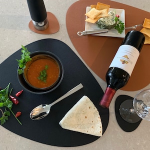 Peut inclure: Un dessous de plat noir avec un bol de soupe de lentilles rouges, un pain plat, une cuillère et des herbes fraîches. Un dessous de plat brun avec une assiette de craquelins et de fromage, une bouteille de vin rouge et un verre à vin.