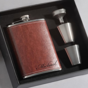 Pode incluir: Uma petaca de aço inoxidável revestida em couro marrom com um funil prateado e dois copos de shot em uma caixa de presente preta. A petaca é gravada com o nome "Michael".