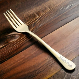 Puede incluir: Un tenedor dorado con cuatro púas, sobre una superficie de madera oscura. El mango del tenedor está grabado con el texto "Alex's Fork". La superficie de madera tiene un tono marrón cálido y rico con vetas visibles.