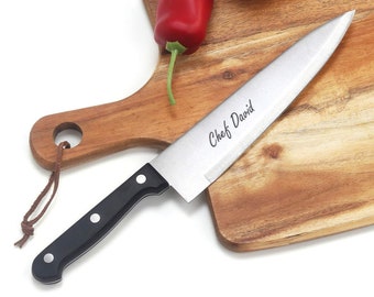 Puede incluir: Un cuchillo de cocina de acero inoxidable con mango negro y las palabras "Chef David" grabadas en la hoja. El cuchillo está sobre una tabla de cortar de madera con dos pimientos rojos.