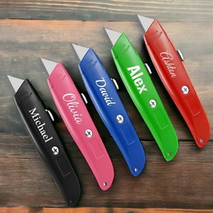 May include: Five utility knives in black, pink, blue, green, and red. Each knife has a silver blade and a name printed on the handle: Michael, Olivia, David, Alex, and Ashton. The text "Warning Sharp Blade" is also printed on each knife.