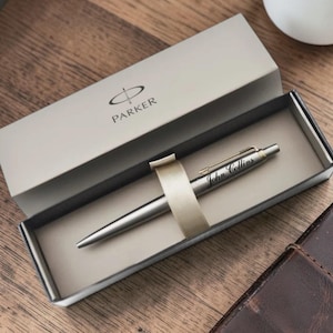 May include: A silver Parker pen with the name "John Collins" engraved on it, presented in a gray box with the Parker logo. The pen is displayed on a wooden surface, next to a cup of coffee and a leather notebook.