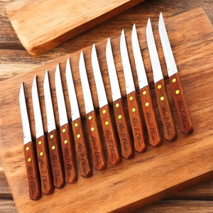 May include: A set of 12 personalised steak knives with wooden handles. The knives are arranged on a wooden chopping board. Each knife has the text "Alex & Olivia" engraved on the handle.